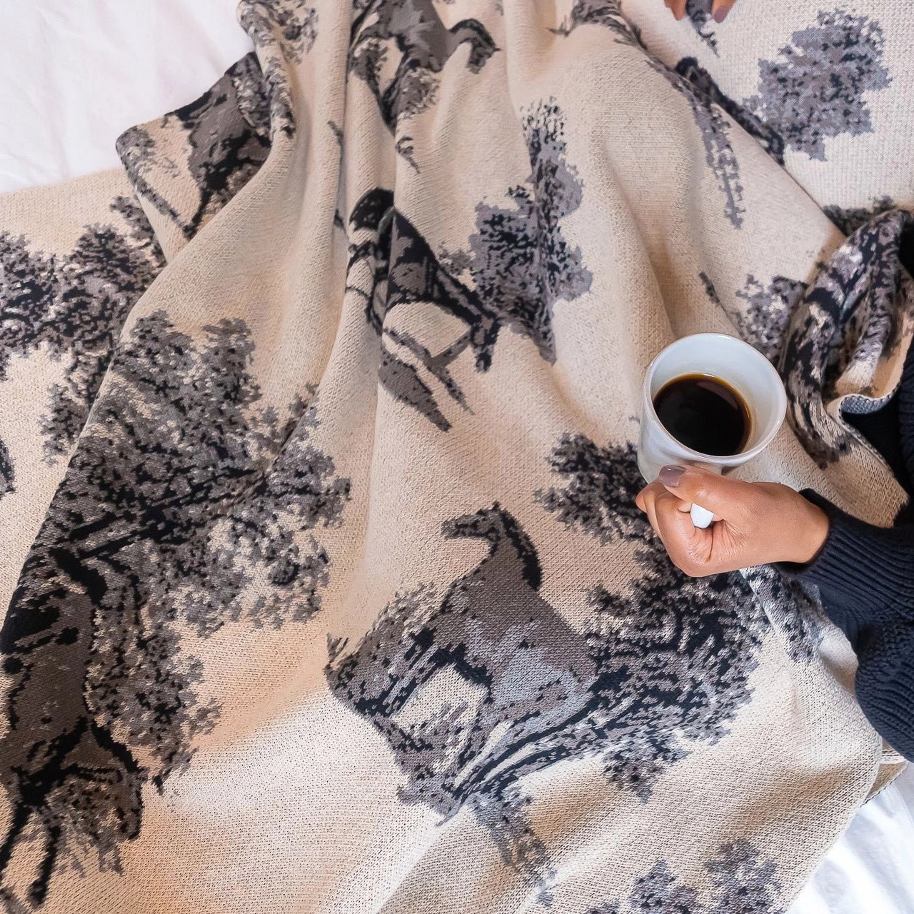 Equestrian Toile Throw Blanket in Charcoal on a women's lap while she drinks a cup of coffee