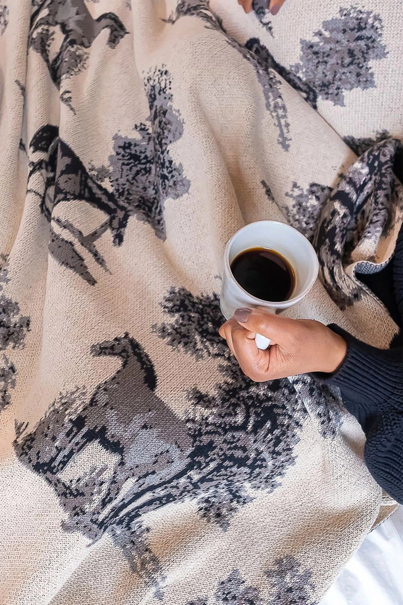 Equestrian Toile Throw Blanket in Charcoal on a women's lap while she drinks a cup of coffee