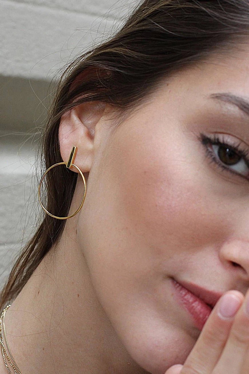 A model is wearing the España Stick Circle Dangle Earrings and her left ear is shown in the image. The vertical post lays on the ear with the circle hanging beneath the ear.