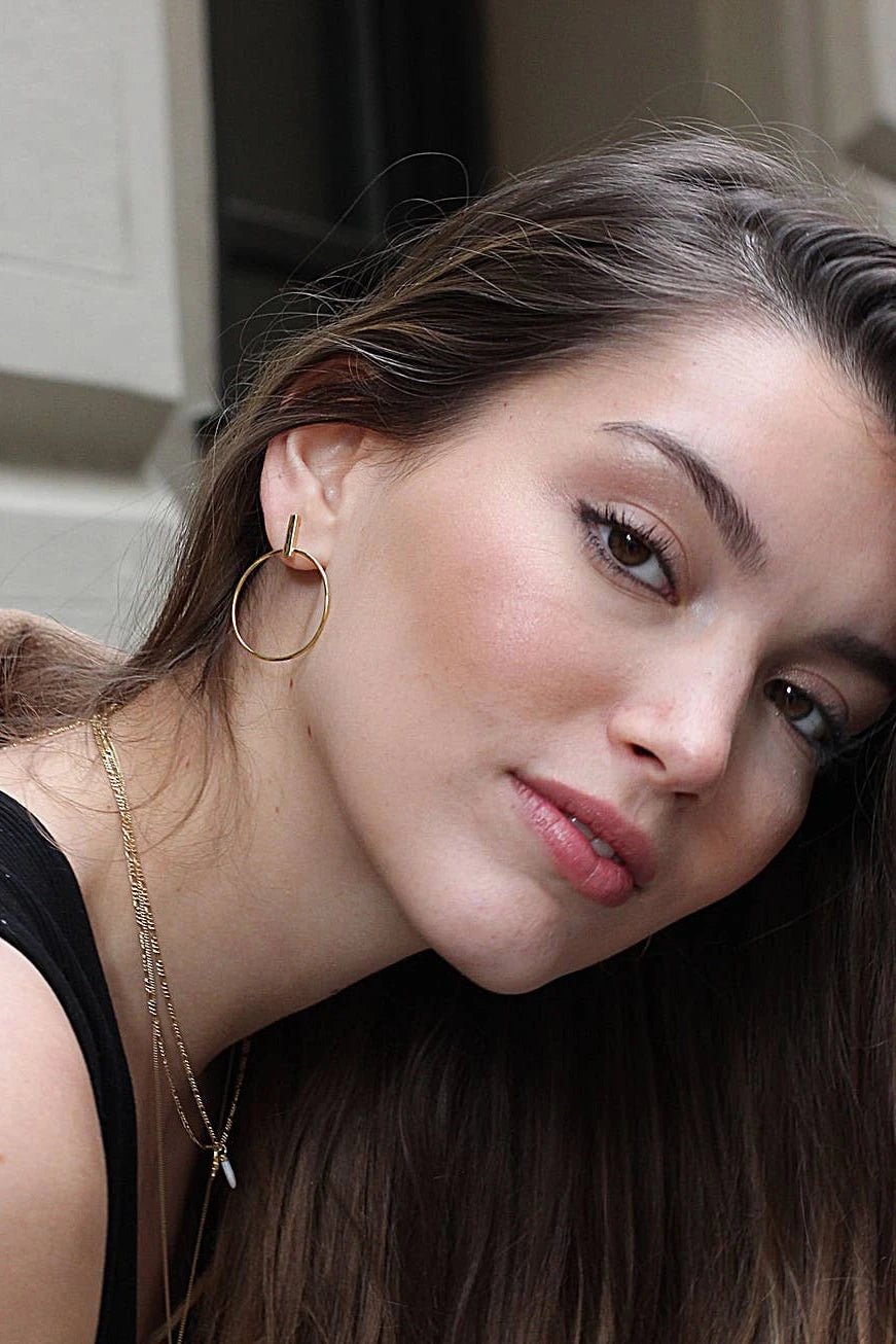 A model is wearing the España Stick Circle Dangle Earrings and her entire face and right ear is shown in the image. The vertical post lays on the ear with the circle hanging beneath the ear