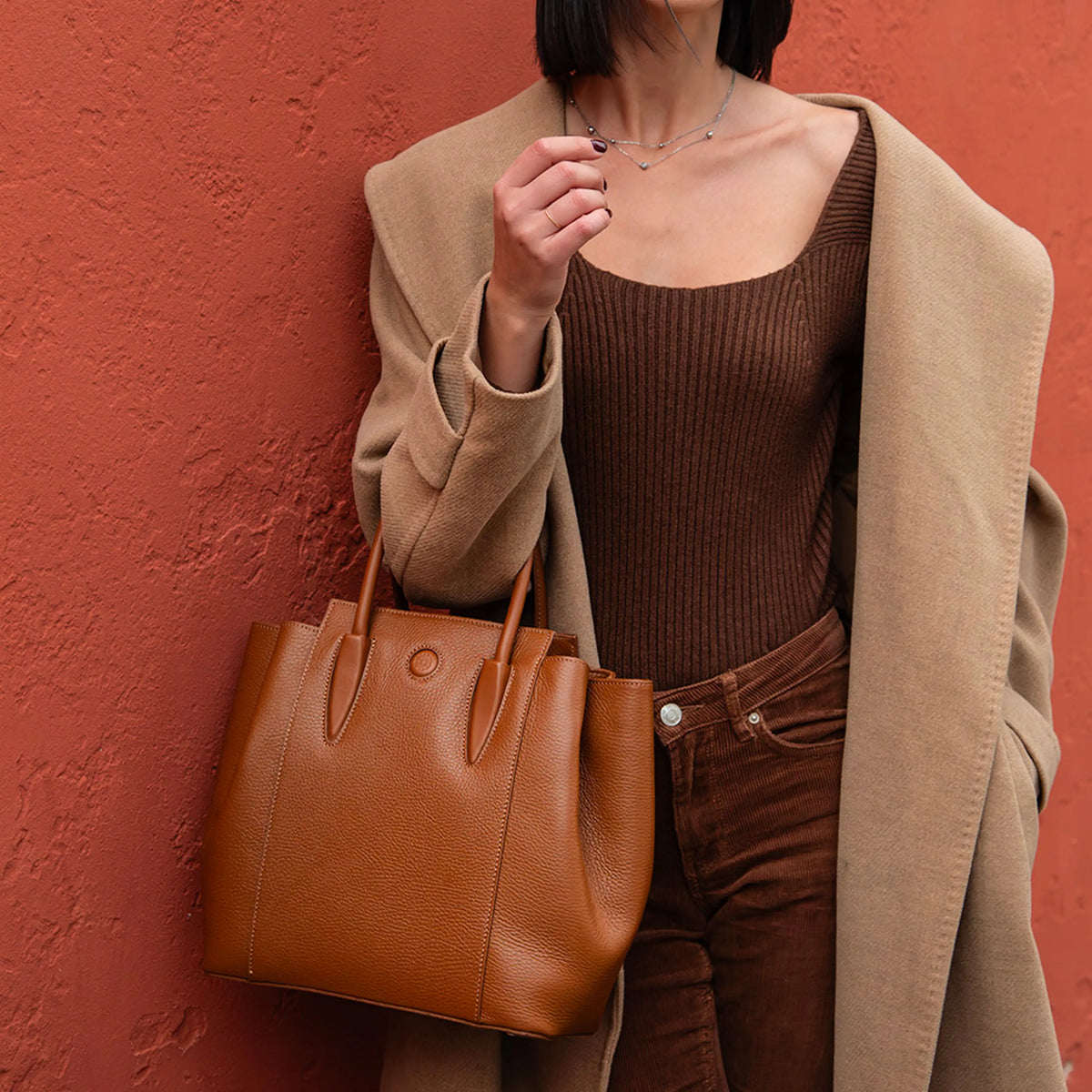 Person wearing a brown outfit with a tan coat and holding the Tulipan cognac brown handbag against a red wall.