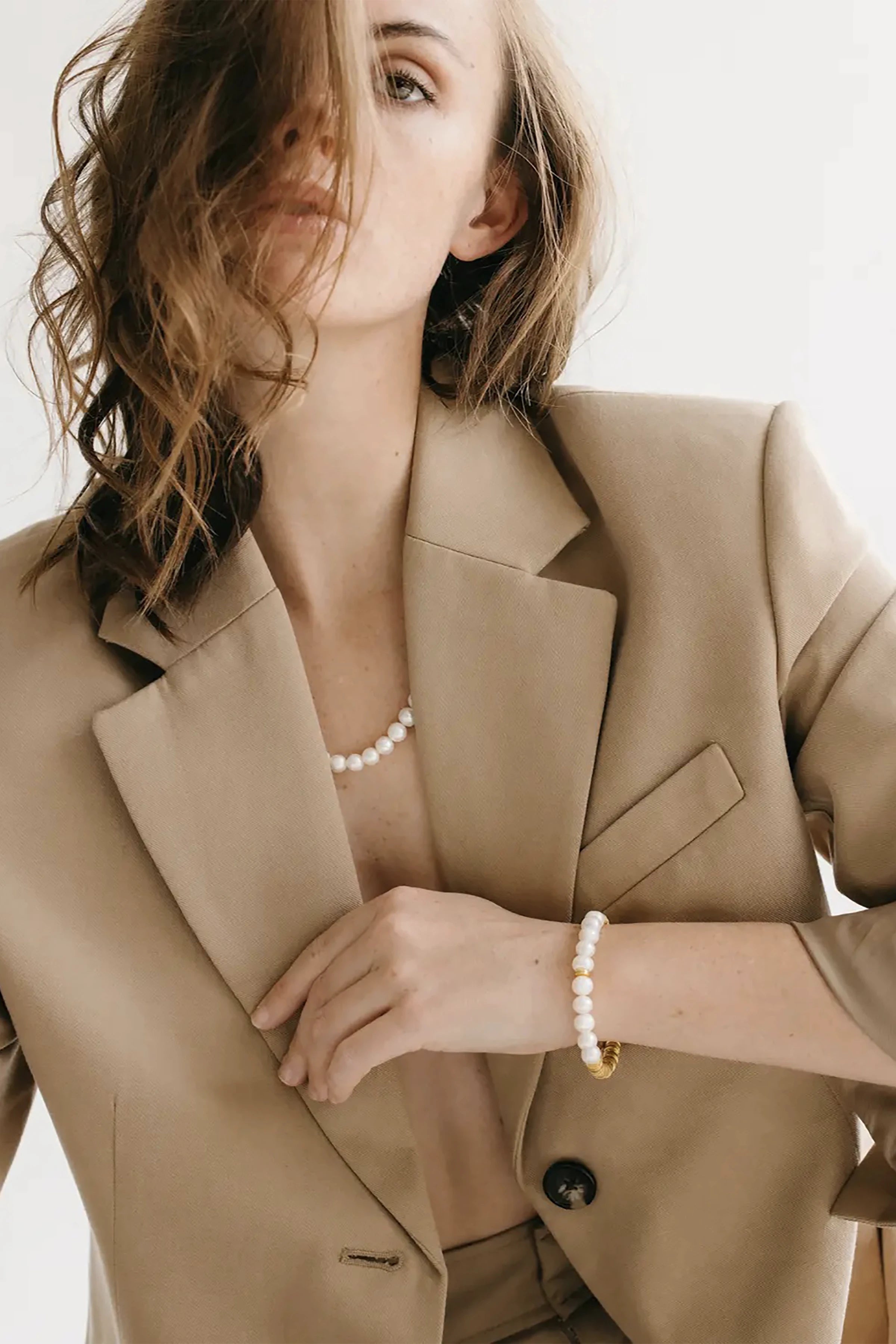 Woman in a beige suit wearing a pearl necklace and pearl with gold bracelet