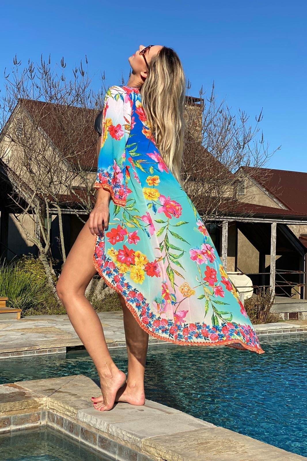 A woman standing by the pool looking left and showing the side and back of the duster. Vibrant flowers on a turquoise