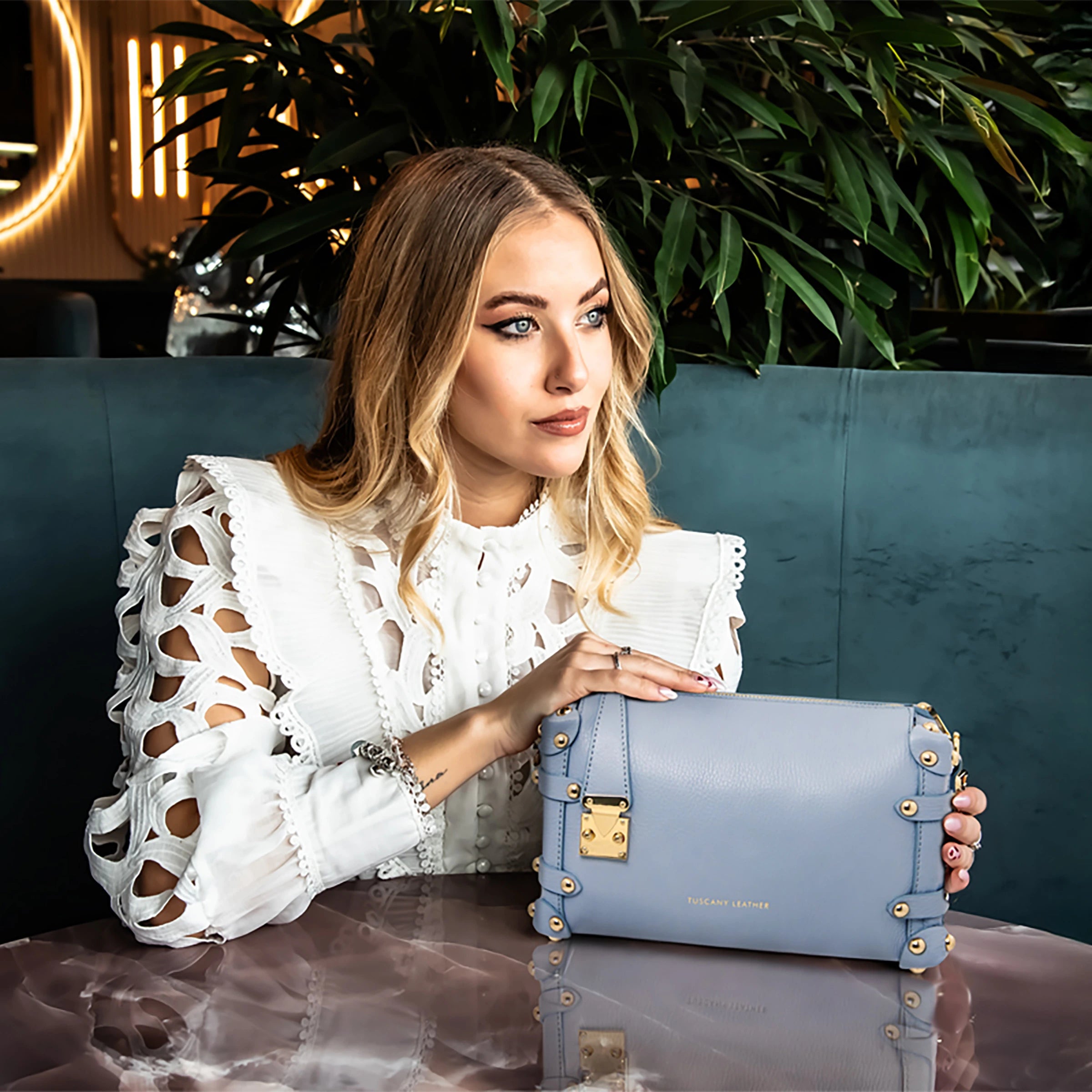 A woman is sitting in a blue leather booth, wearing a cut all over out white shirt, holding the Praline bag in light blue on a wooden table