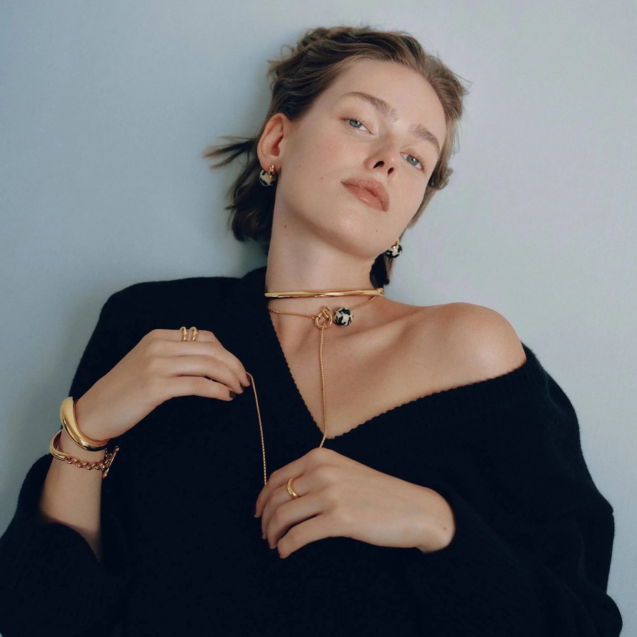Front view of Golden Arc Collar Necklace on a woman's wearing all gold jewelry leaning on a light blue wall