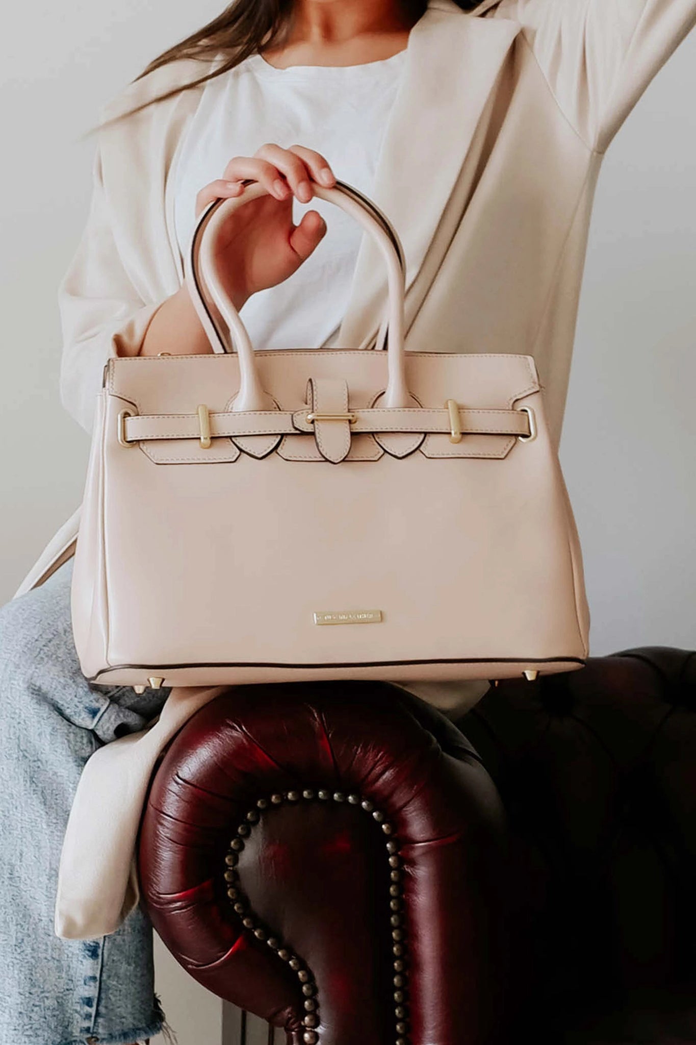 Model sitting on brown leather couch arm holding the TL Bag Italian Leather Handbag in Nude by the handle. Bag is set on the arm.