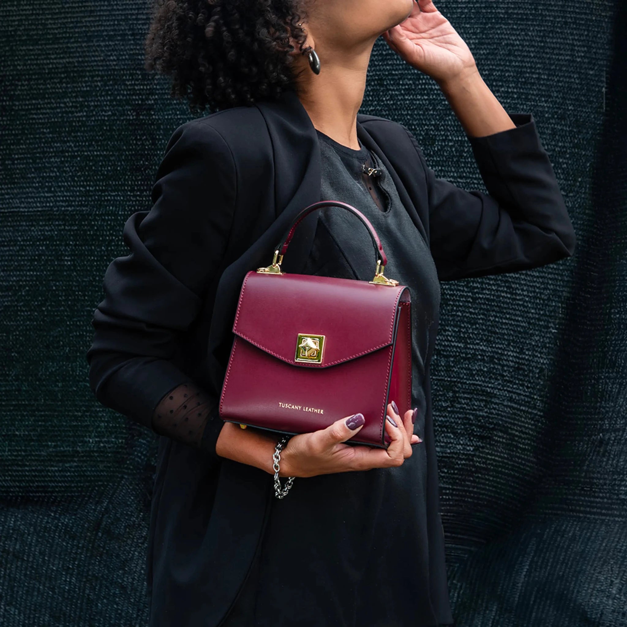Person holding a bordeaux hued handbag with a visible brand logo against a dark textured background
