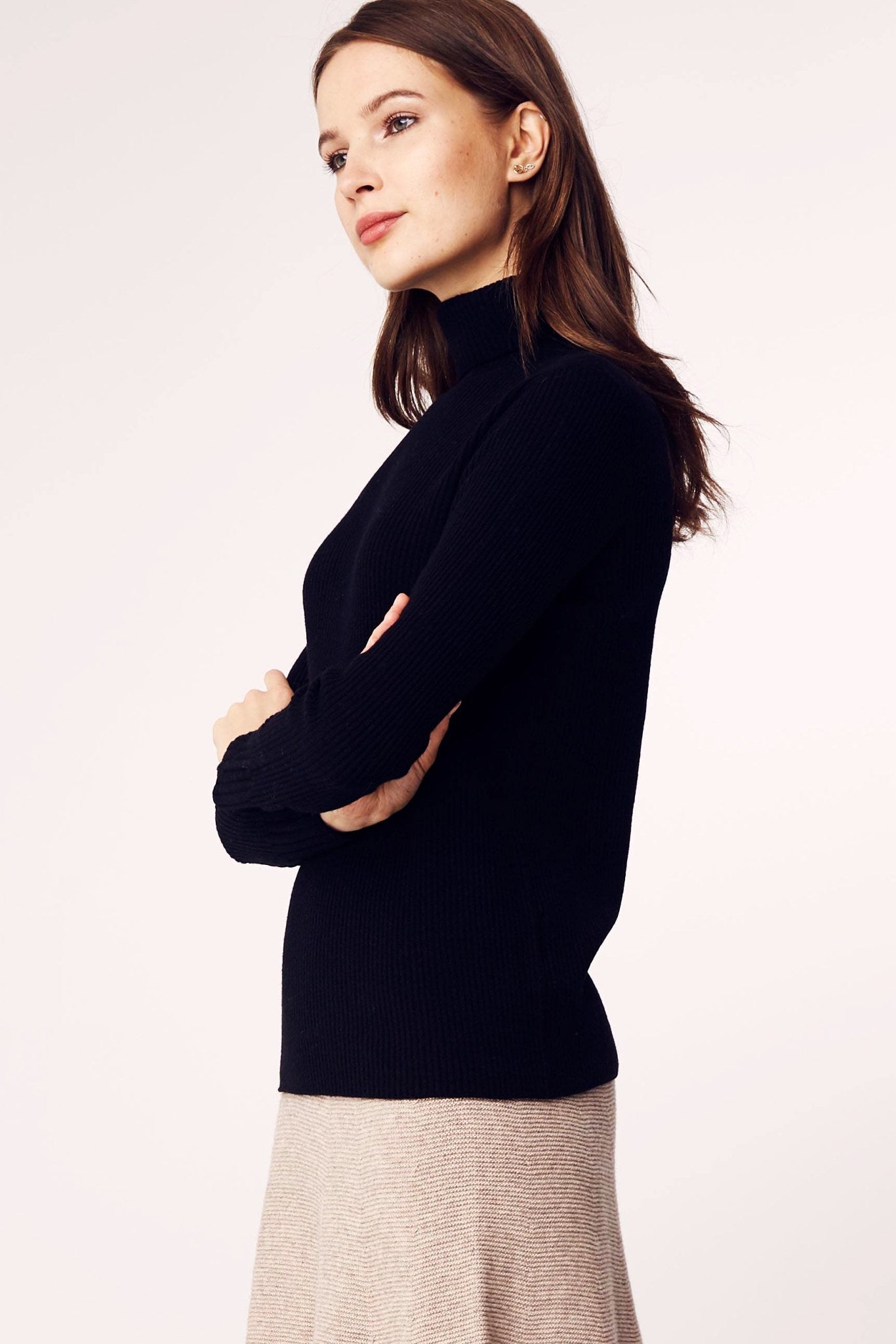 Model is standing and wearing the Seamless Ribbed Classic Turtleneck Sweater in Black on a khaki mid length skirt