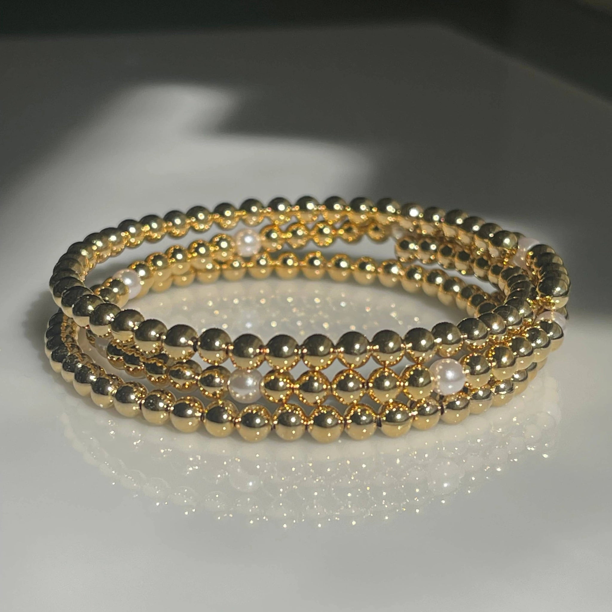 Set of 3 golden beaded bracelets with pearls laying on a glass table showing a reflection
