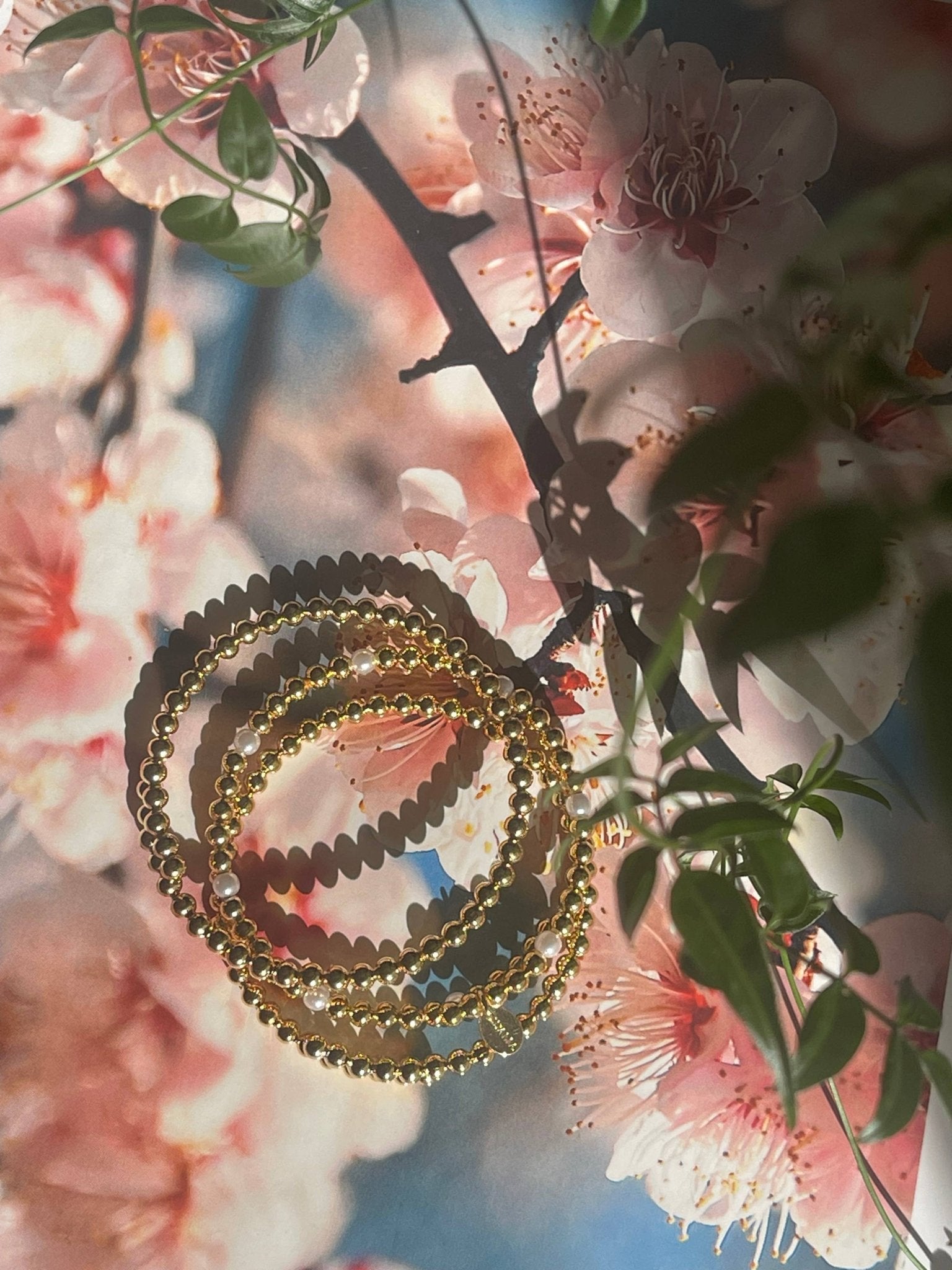 Set of 3 golden beaded bracelets with pearls laying on desk on a pink floral background