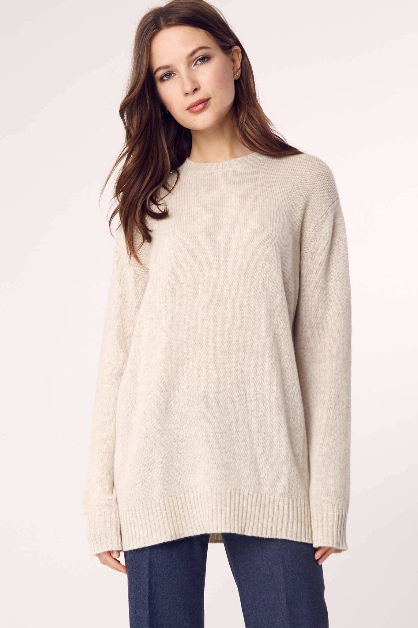 A woman is wearing the Cashmere Classic Long Crew Sweater in Sand on blue pants and standing in front of a white background