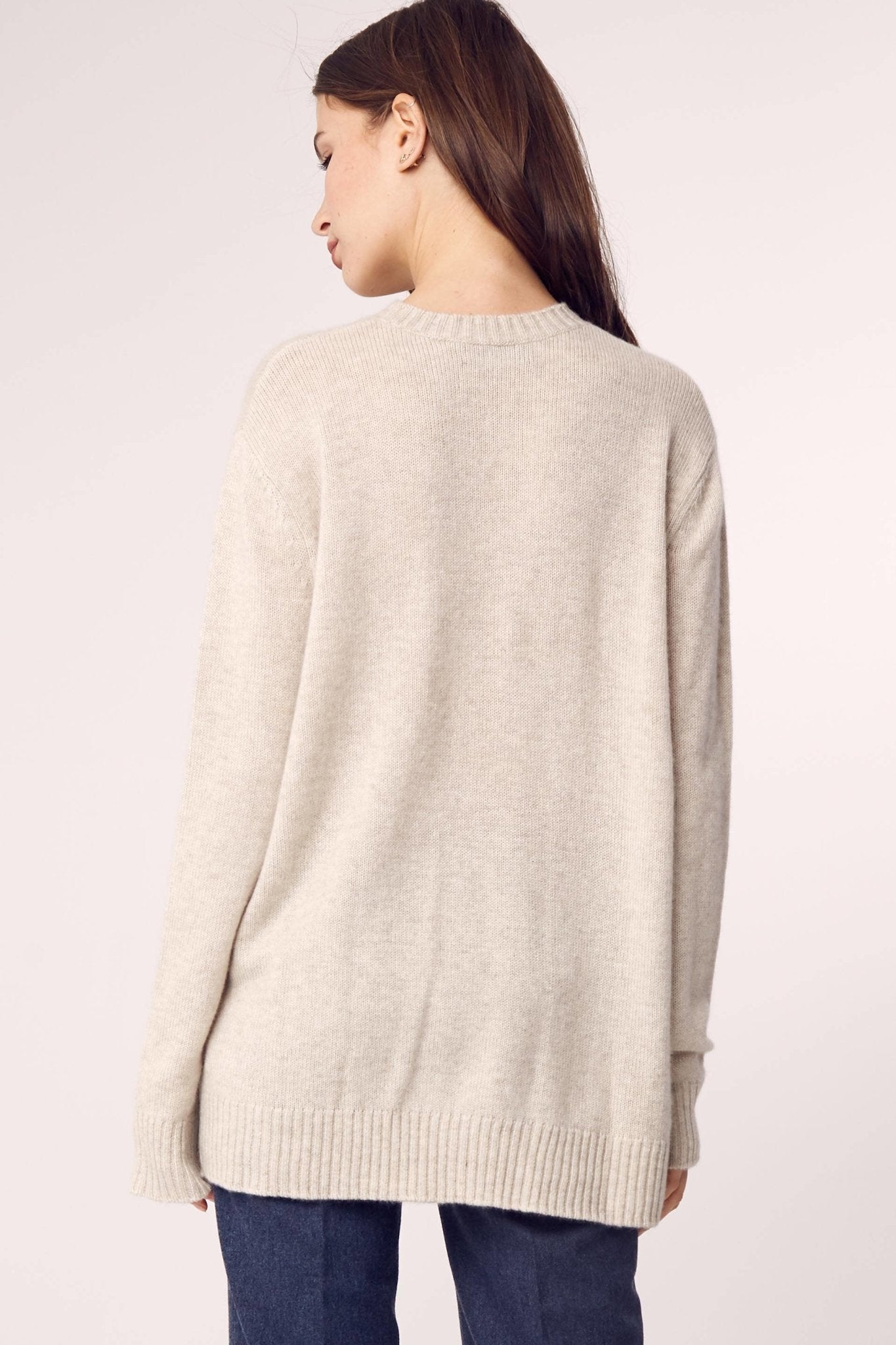 A woman is wearing the Cashmere Classic Long Crew Sweater in Sand on blue pants and is showing the back of the below the hips garment