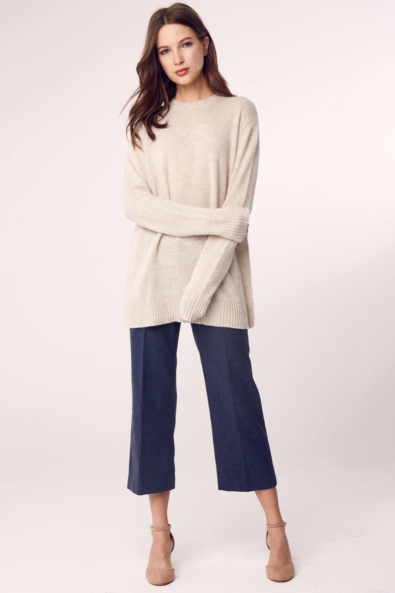 A woman is wearing the Cashmere Classic Long Crew Sweater in Sand on blue capri pants and standing in front of a white background