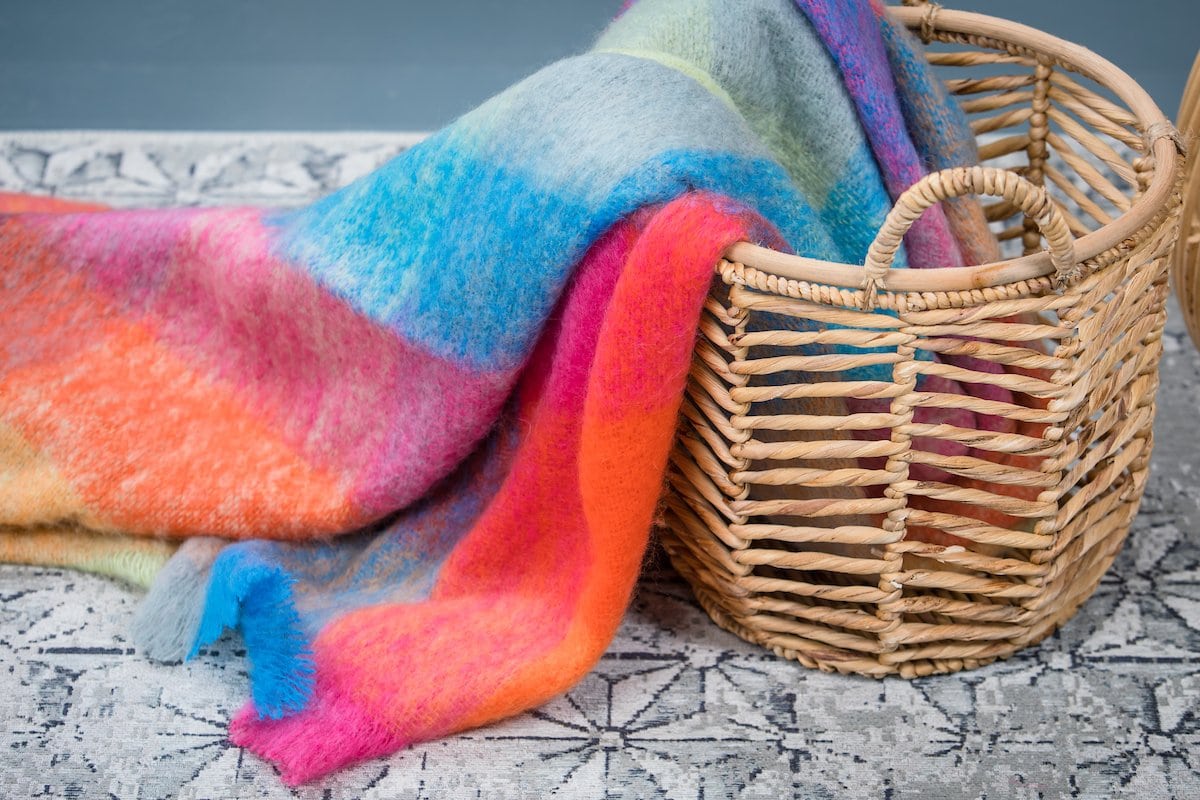 Color Block Luxuriously Soft Irish Mohair Throw laying partially inside a wicker basket and a black and white tile floor