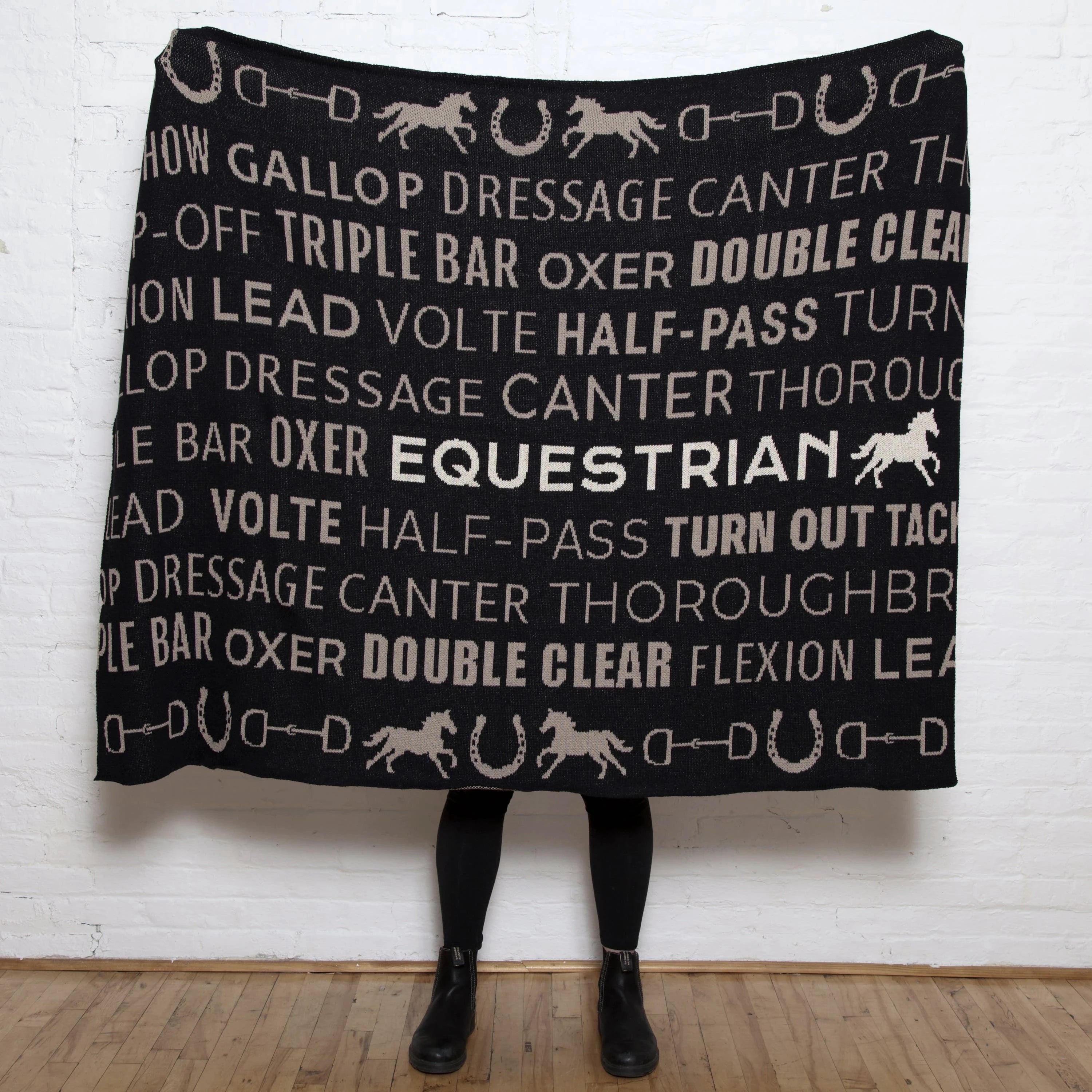 A woman is holding the Equestrian Eco Text Throw fully opened in front of her and all you see are her black boots.