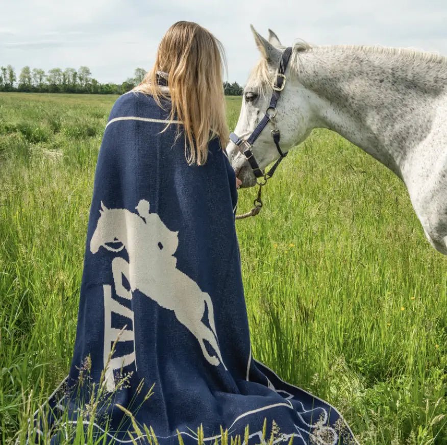 A woman is standing in a grassy field wrapped in the Equestrian Jumper Reversible Throw and petting a white thoroughbred