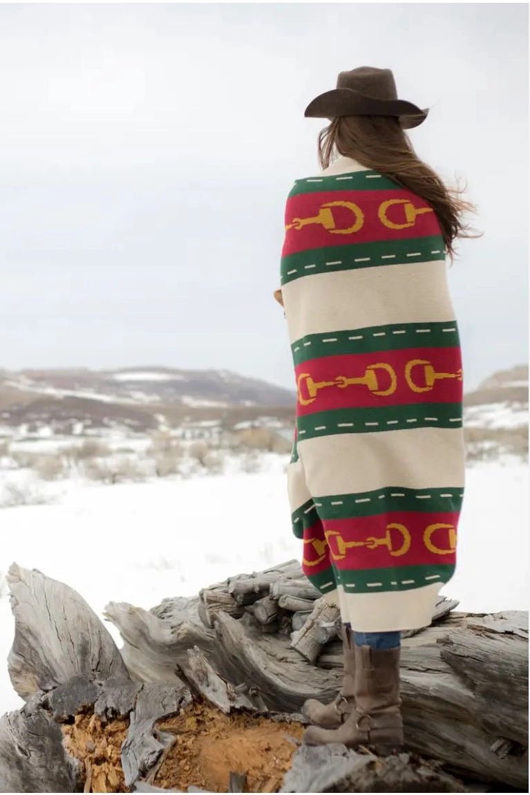 A woman is standing o nboulders staring at a snowy field wrapped in the Equestrian Striped Horse Bit Eco Throw and wearing a brown cowboy hat.