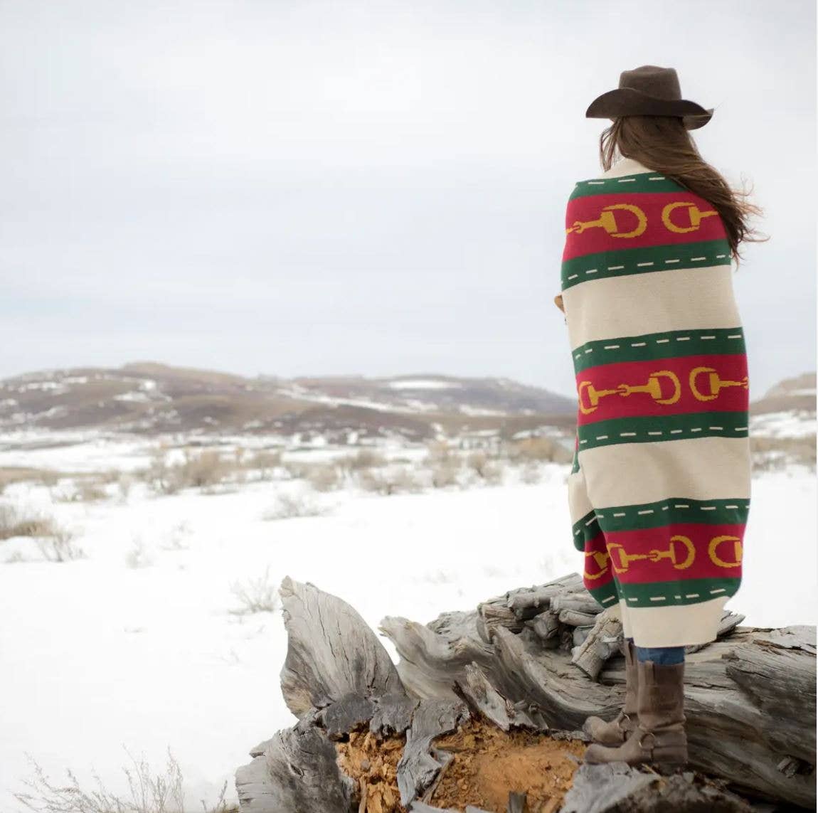 A woman is standing o nboulders staring at a snowy field wrapped in the Equestrian Striped Horse Bit Eco Throw and wearing a brown cowboy hat.