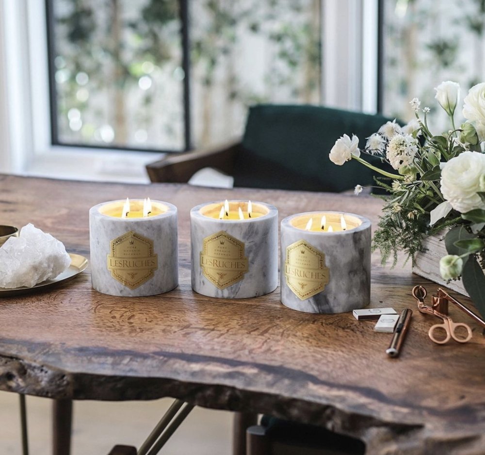 Three Les Ruches 32 oz candles on a wooden table next to a white floral bouquet