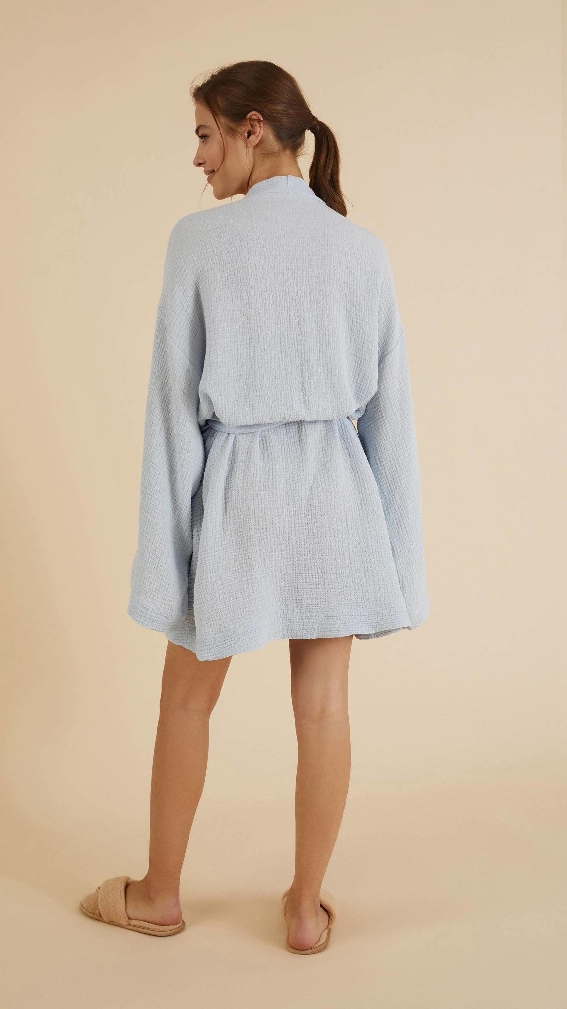 Luxe Luna Kimono Wrap in baby blue on a model in front of a natural-hued background with light. She is presenting the back of the robe.