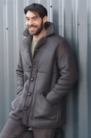 A man wearing a gray coat with his hands in his pockets is standing in front of a metal wall.