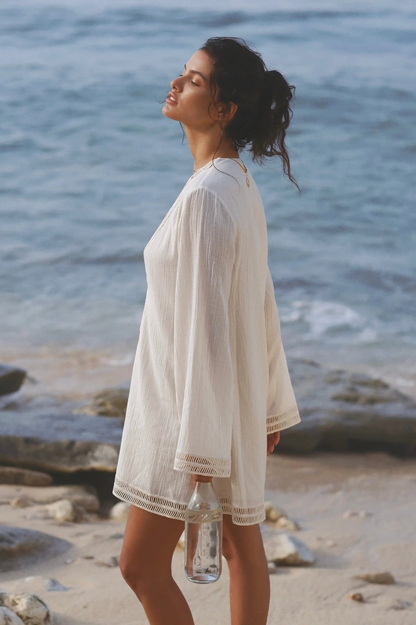 Positano Turkish Cotton Cover Up Dress with Lace in Natural on model standing on on the beach holding a glass water bottle.