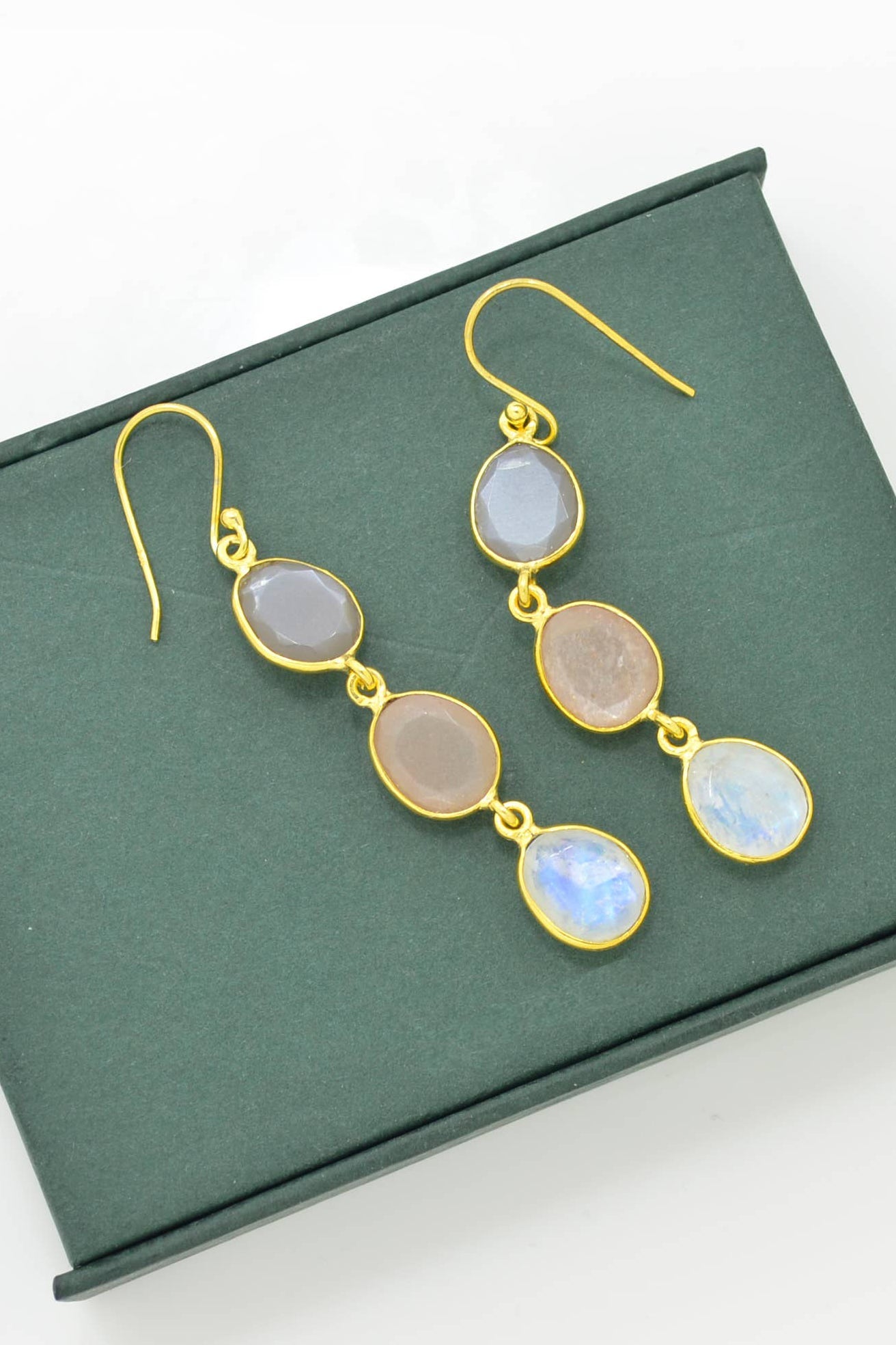 Rainbow, Grey & Peach Moonstone Earrings laying on a forest green background showcasing the earthy hues of the moonstone.