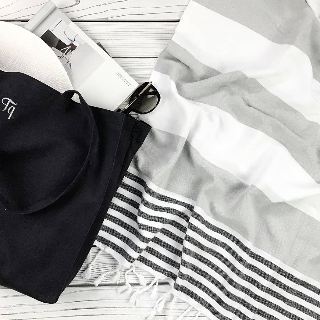 Riviera Turkish Cotton Gray and White Striped Towel in Gray laying on a light gray washed deck next to a black beach bag and sunglasses to the left