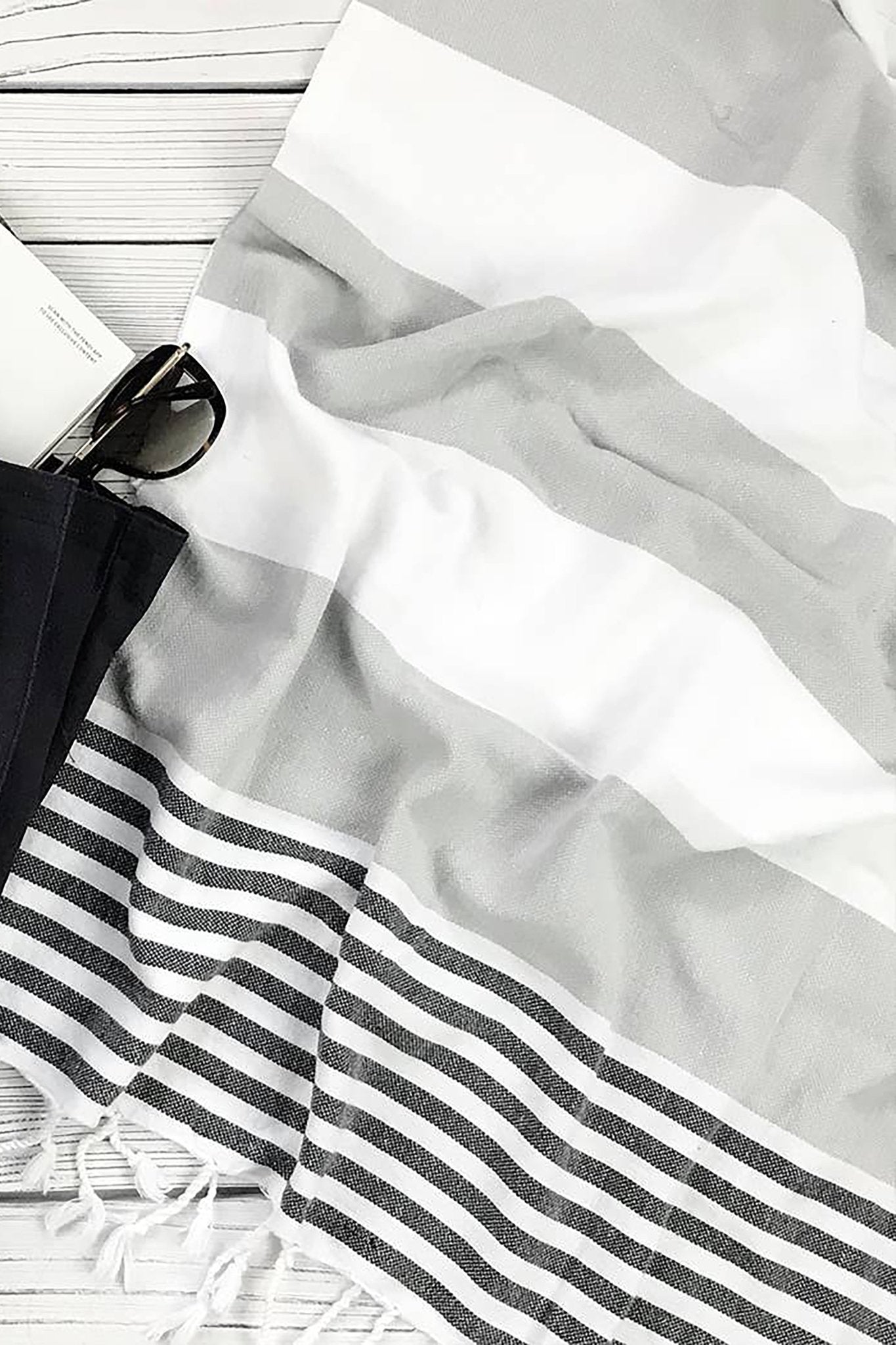 Riviera Turkish Cotton Gray and White Striped Towel in Gray laying on a light gray washed deck next to a black beach bag and sunglasses to the left