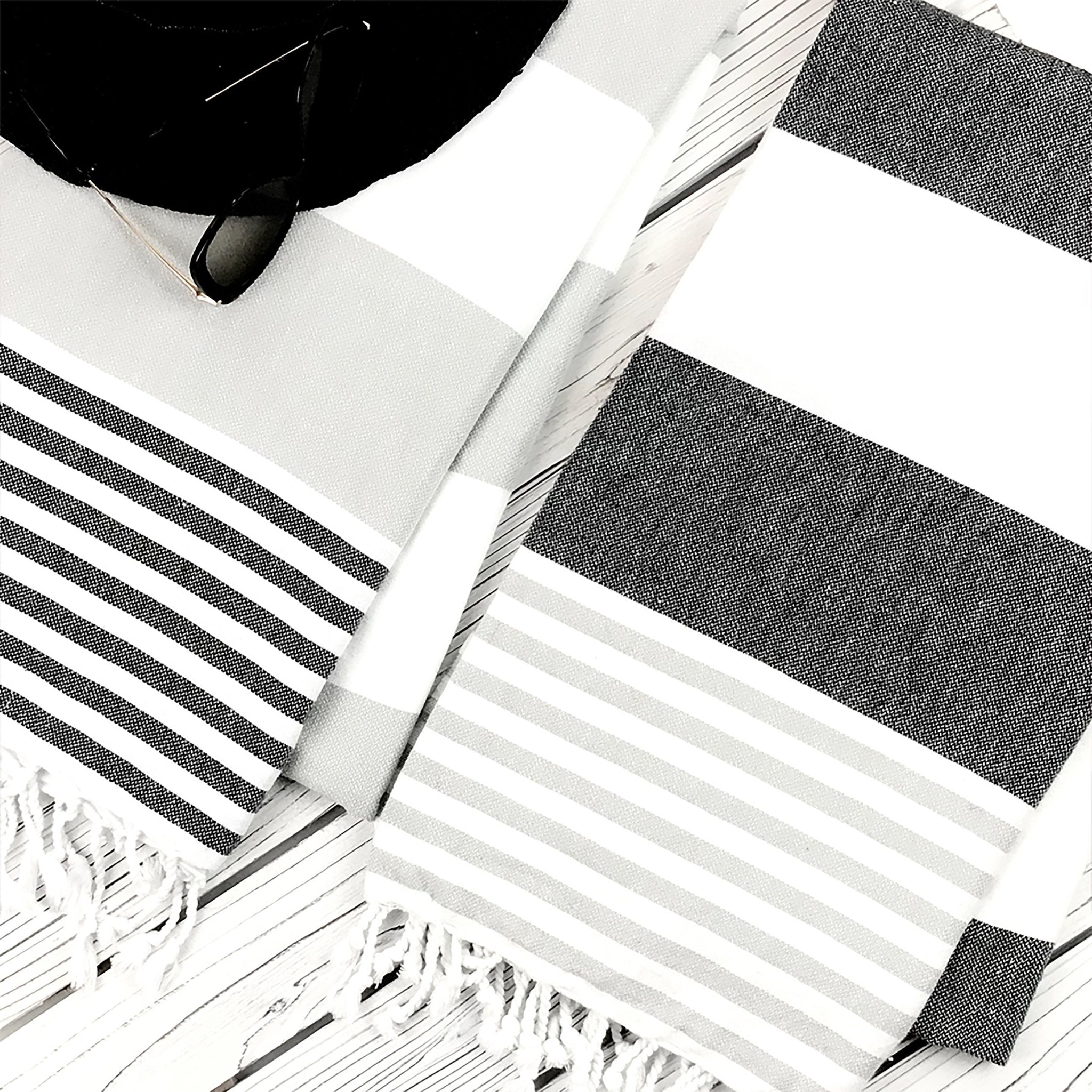 Riviera Turkish Cotton Gray and White Striped Towel in Gray and Black laying on a light gray washed deck next to a black black beach hat and sunglasses on the upper left.
