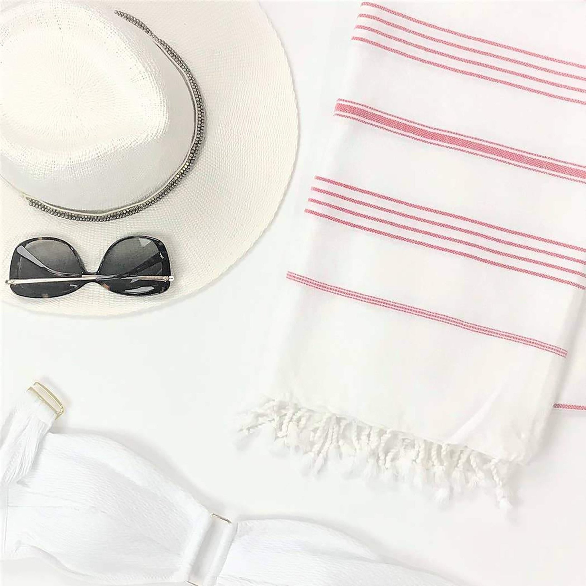 Signature Turkish Cotton Striped Towel in Pink on a white background with a straw sun hat, white bikini strapless top and sunglasses.