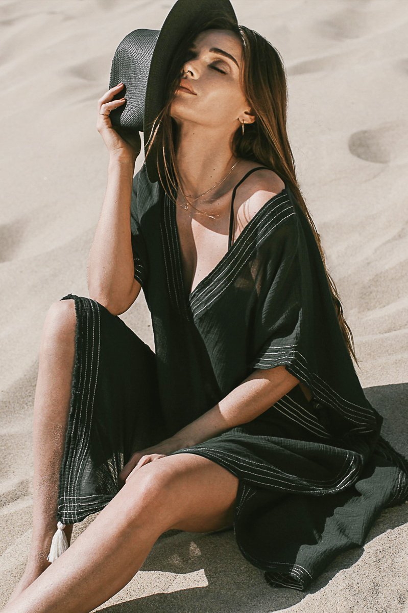 A woman wearing Summer Ready Breezy Kaftan in Black sitting on the beach, holding a black hat.