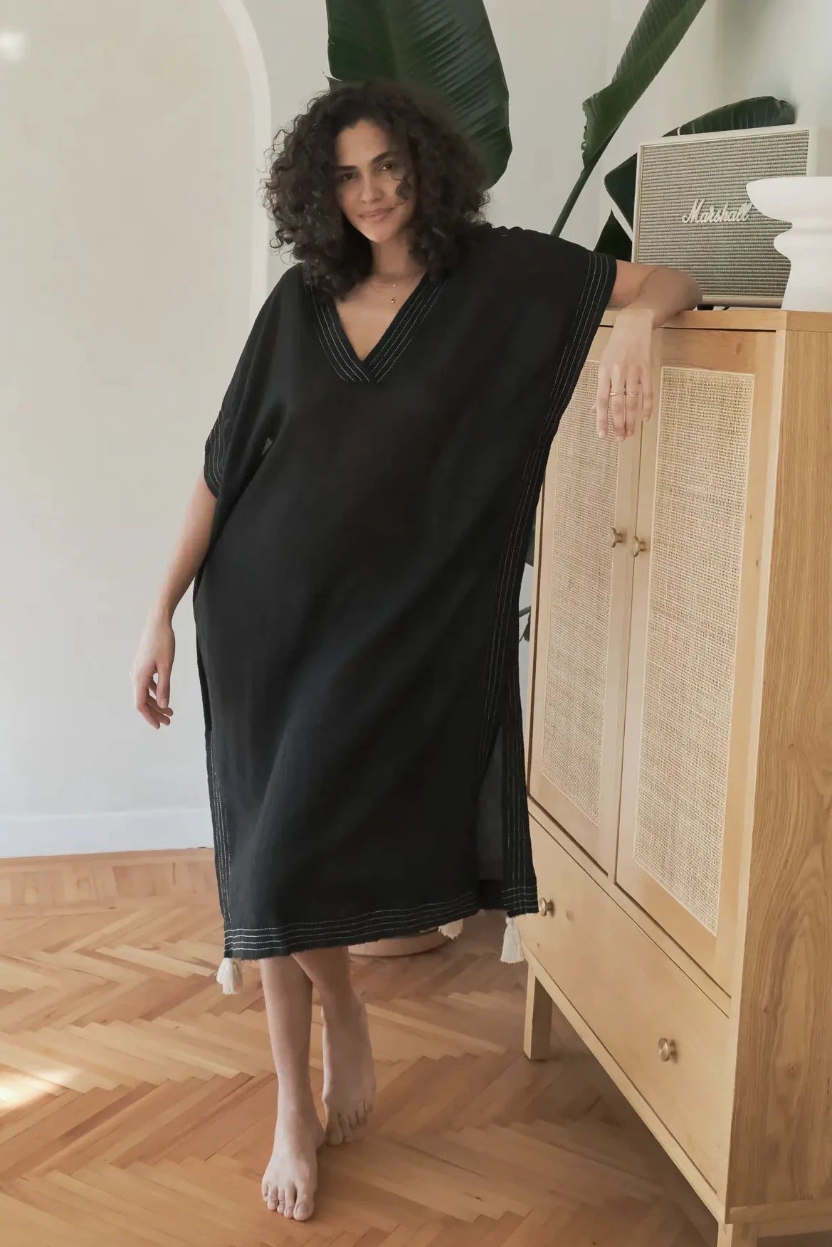 A woman wearing Summer Ready Breezy Kaftan in Black leaning on a cabinet