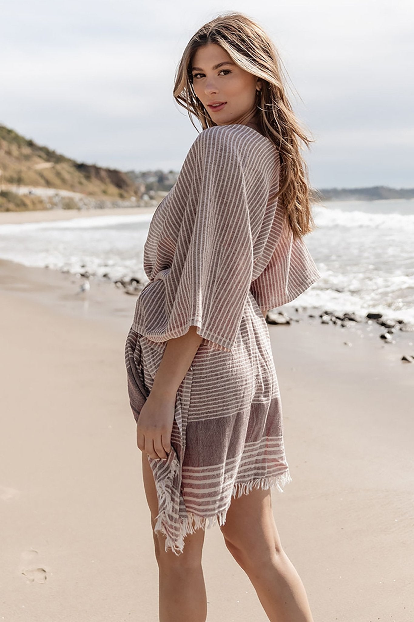 Turkish Aegean Cotton Striped Kimono in Dark Cherry on model standing on a Turkish beach. She is presenting the side and back of the cover up.