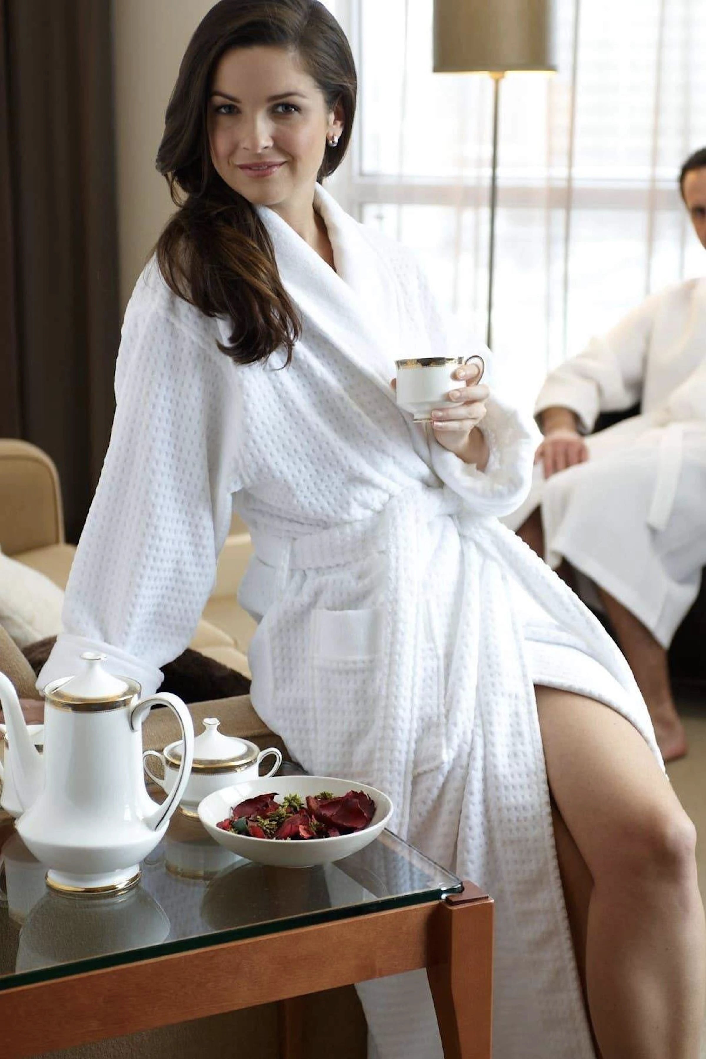 A female model is wearing the Unisex Jacquard Cotton Terry Velour Robe, holding a cup of coffee and sitting on the arm of a couch in a bedroom setting