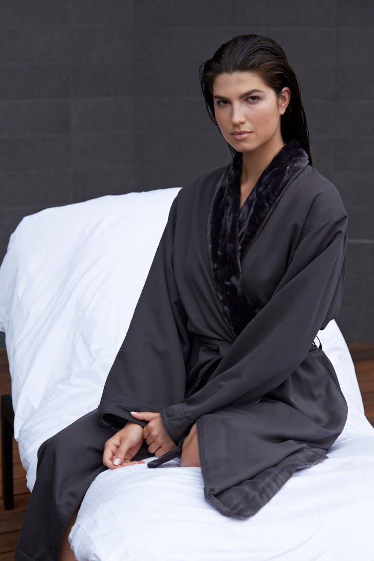 A woman is sitting on a spa lounge wearing the Unisex Spa Microfiber Shimmer Lined Robe in charcoal