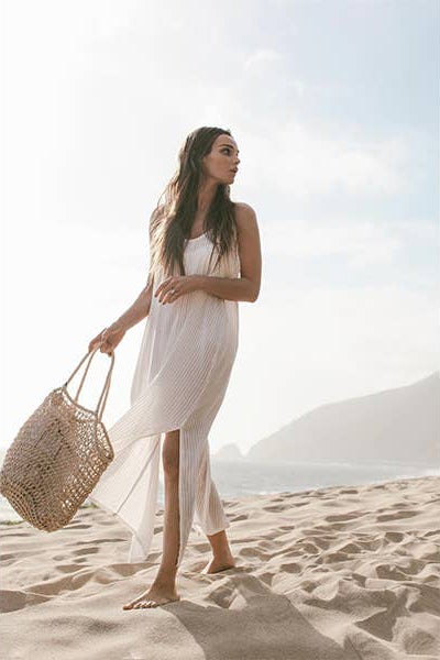 Yaz Dress in Natural with White Stripes on model walking with a crochet tote bag on the beach.