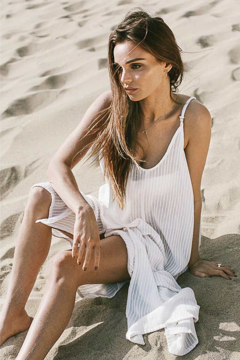 Yaz Dress in Natural with White Stripes on model sitting on the sand on the beach.