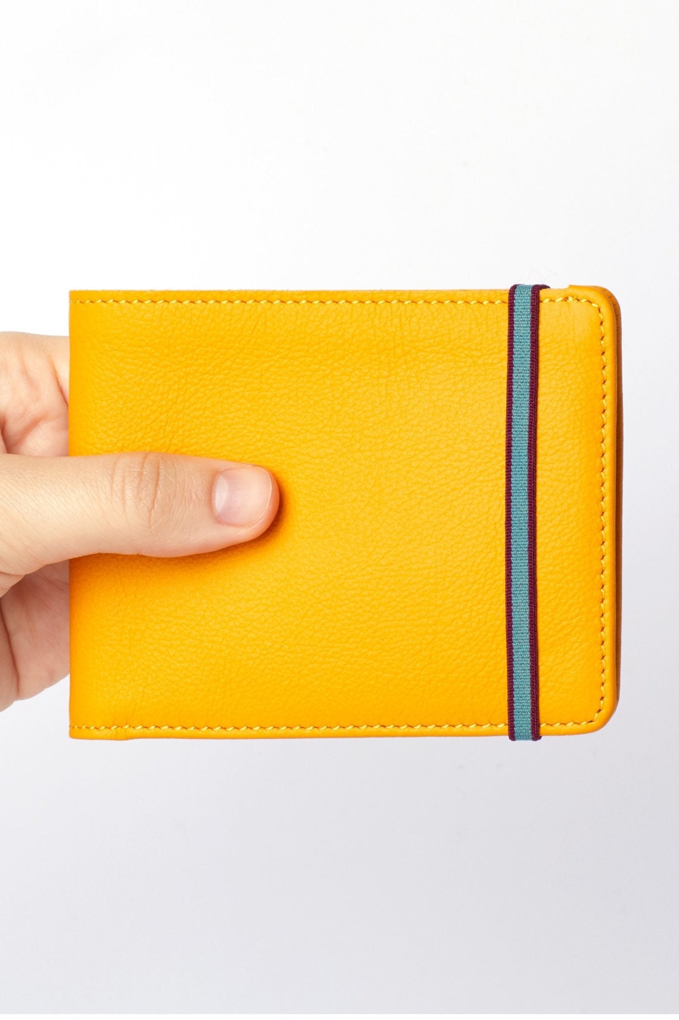 A man's hand holding the Yellow Wallet with a light blue and burgundy elastic closure on white background - L'Atelier Global
