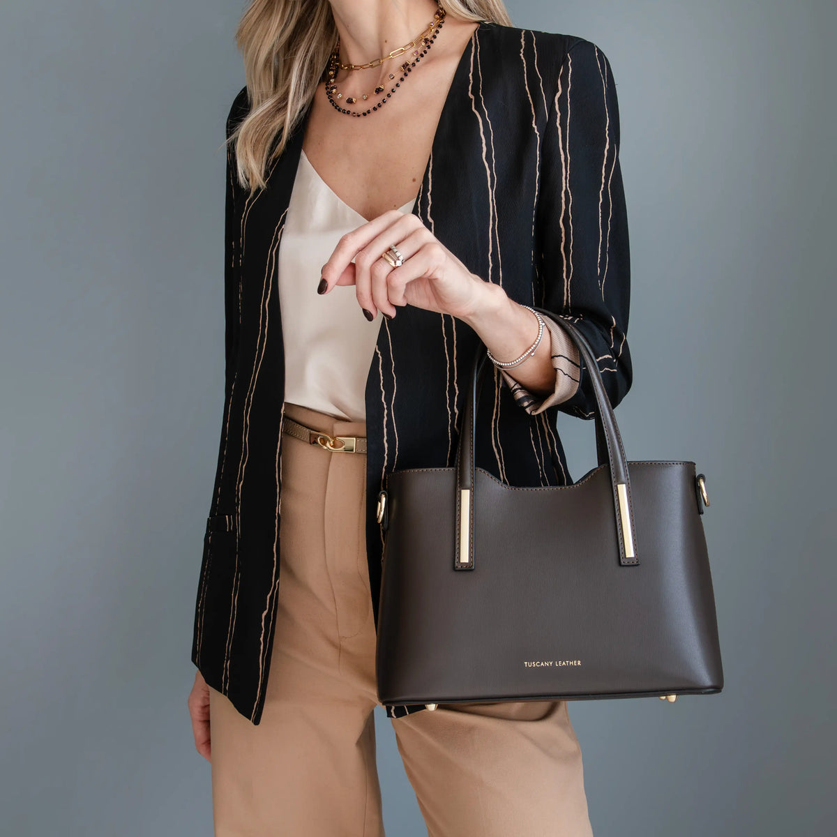A woman models a dark brown structured handbag with gold-tone hardware, styled with a black and beige striped blazer, a cream camisole, and tan trousers against a solid gray studio background.