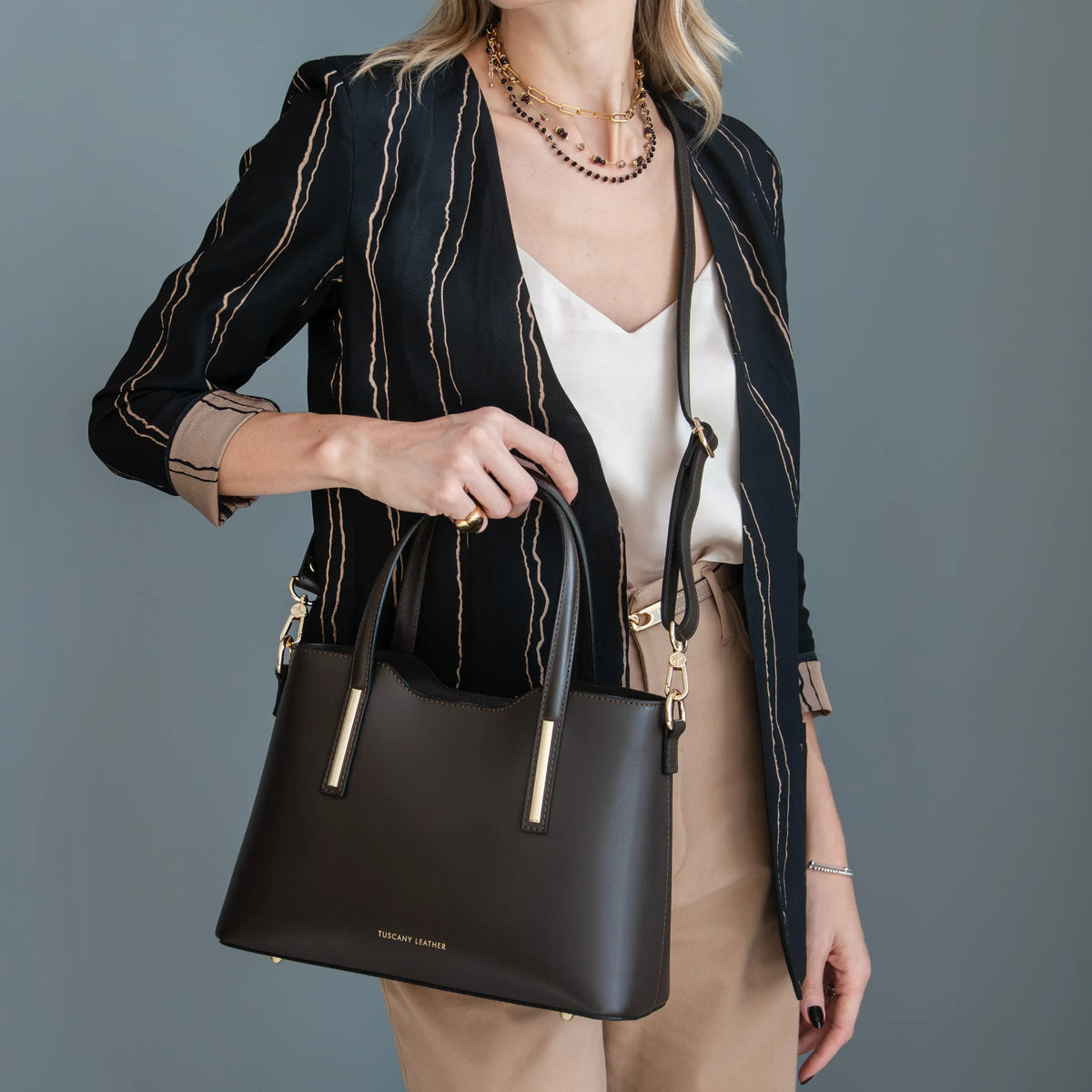 A woman models a dark brown leather handbag with gold-tone hardware and a top handle, styled with a black and beige striped blazer, white top, and beige pants against a solid gray background.