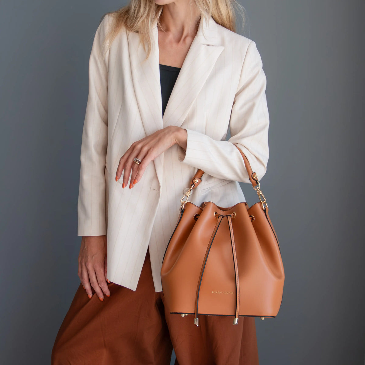 A model wearing a cream blazer and brown trousers carries a cognac leather drawstring bucket bag with gold-tone hardware against a dark gray studio background.