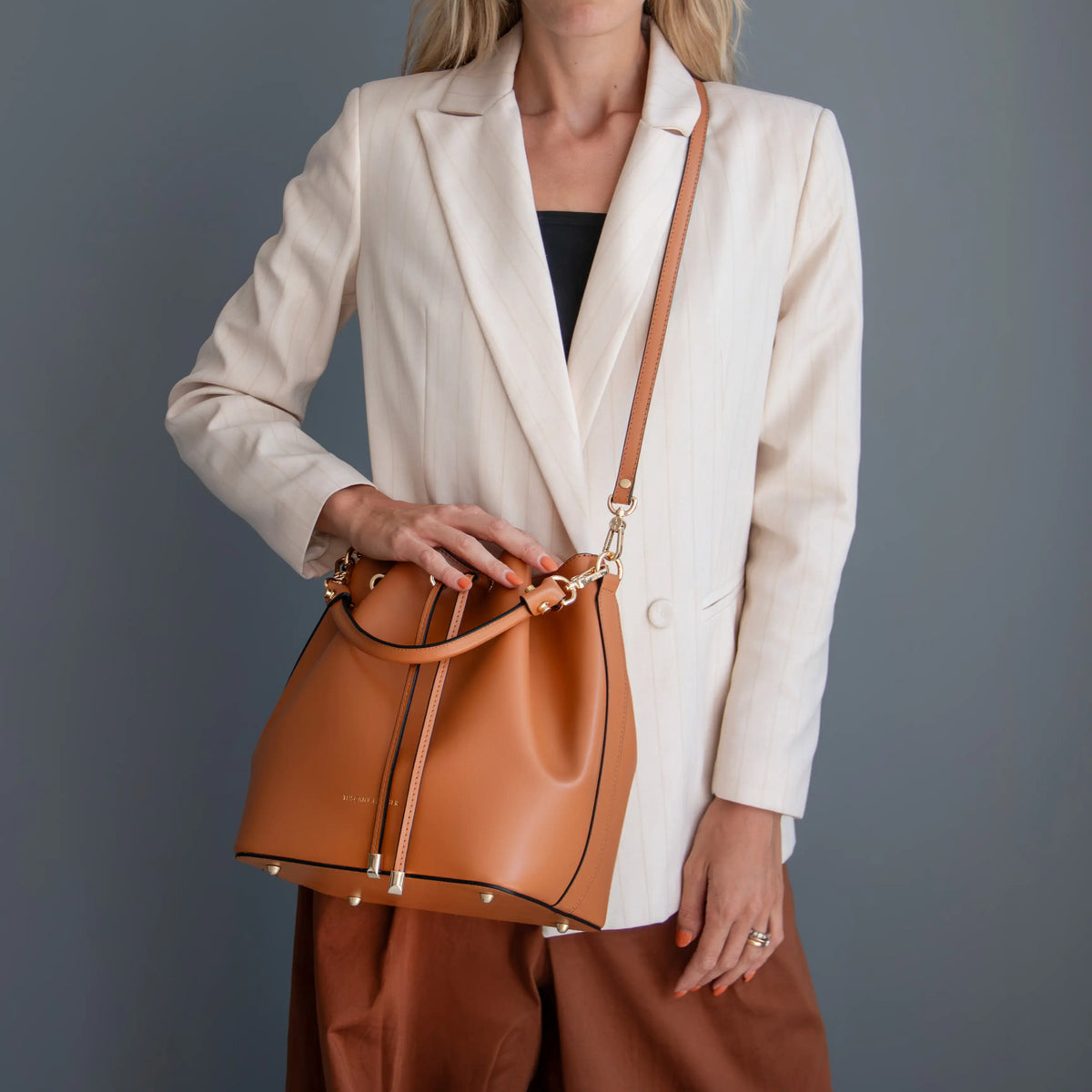 A woman wearing a cream blazer and brown pants carries a cognac leather bucket bag with a matching shoulder strap and gold-tone hardware against a dark gray studio background.