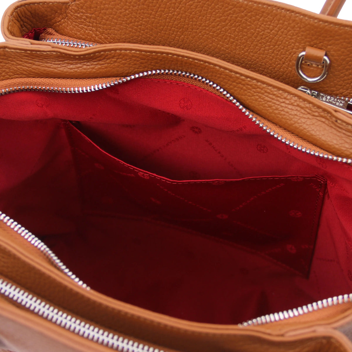 Close-up of a cognac pebbled leather handbag showing the interior open pocket with a bright red fabric lining and silver-tone zippers.