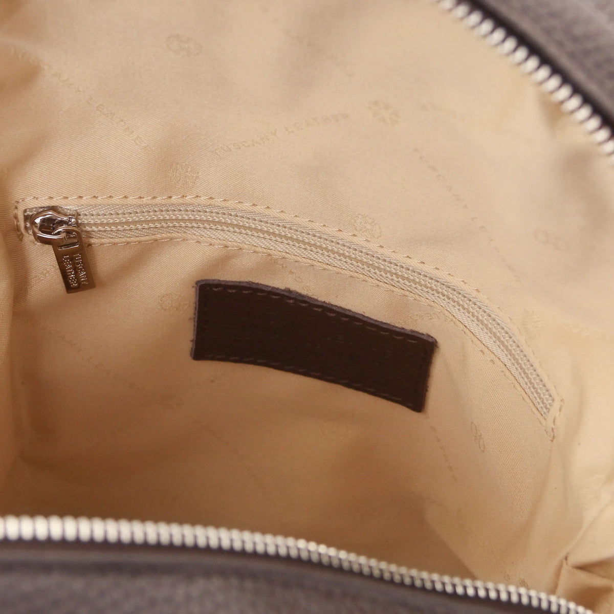 Close-up of the interior of a bag showing a beige quilted lining with a zippered pocket, a dark brown brand patch, and silver-toned metal zipper hardware.