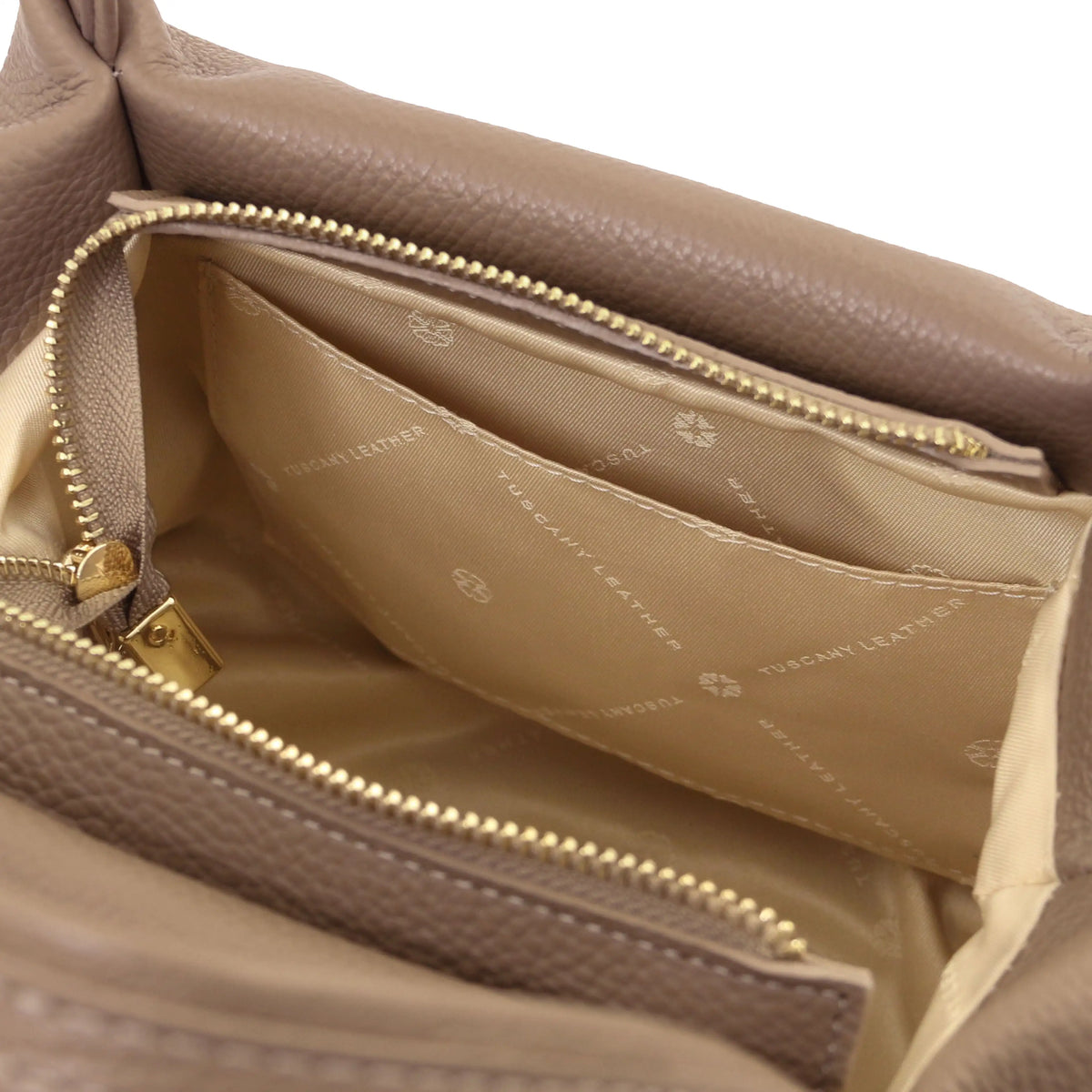 Close-up of the interior of a taupe leather bag, showing a gold-tone zipper, a beige interior open pocket.