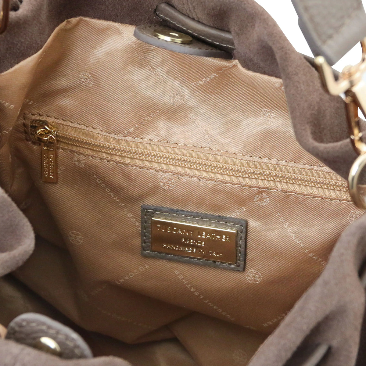 Close-up of the interior of a shoulder bag, showing a beige patterned nylon lining with a gold-tone zipper, a metal brand plaque, and dark taupe suede-like trim.