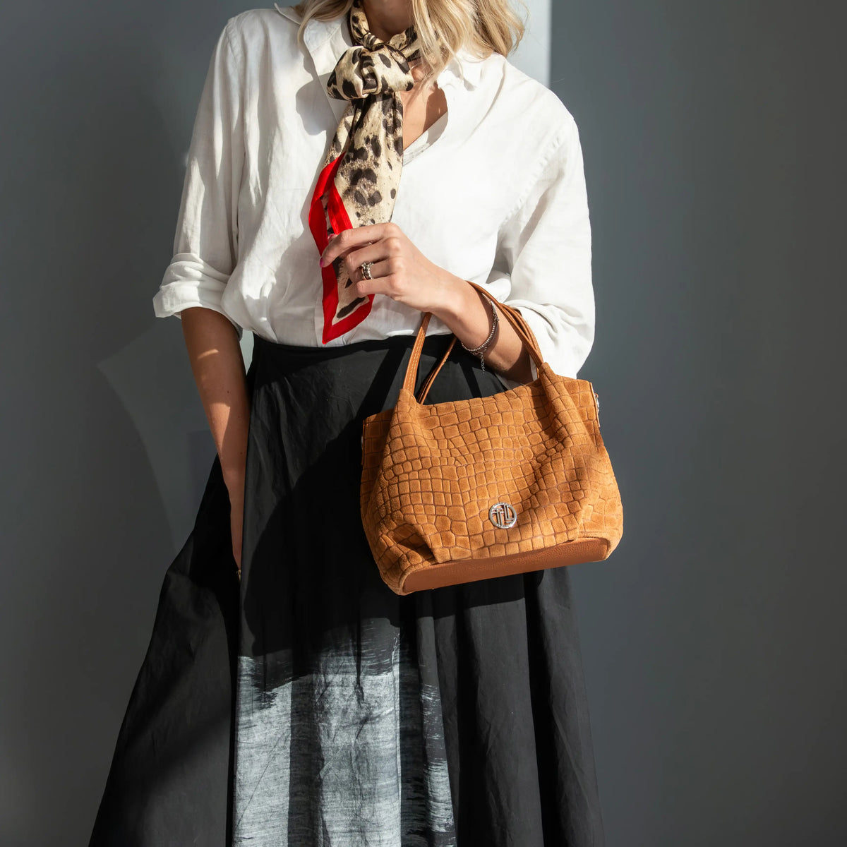 A woman wearing a white blouse, black skirt, and a leopard-print neck scarf holds a cognac crocodile-embossed leather handbag with a silver-tone logo on a dark gray background.