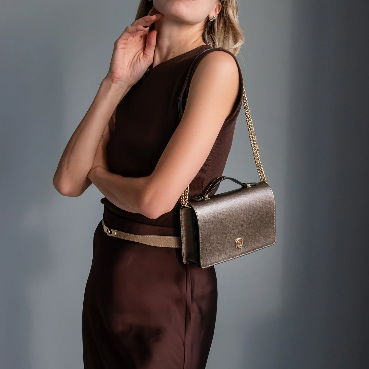 A woman wearing a dark brown sleeveless dress and a beige belt carries a structured metallic bronze handbag with a gold chain strap and top handle against a dark gray studio background.