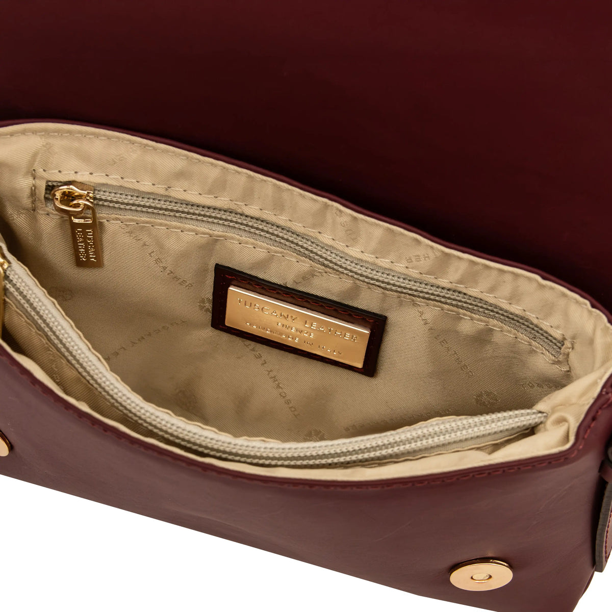 Close-up of the interior of a burgundy leather handbag showing a beige lining and zippered pocket with a gold-tone metal brand plaque.